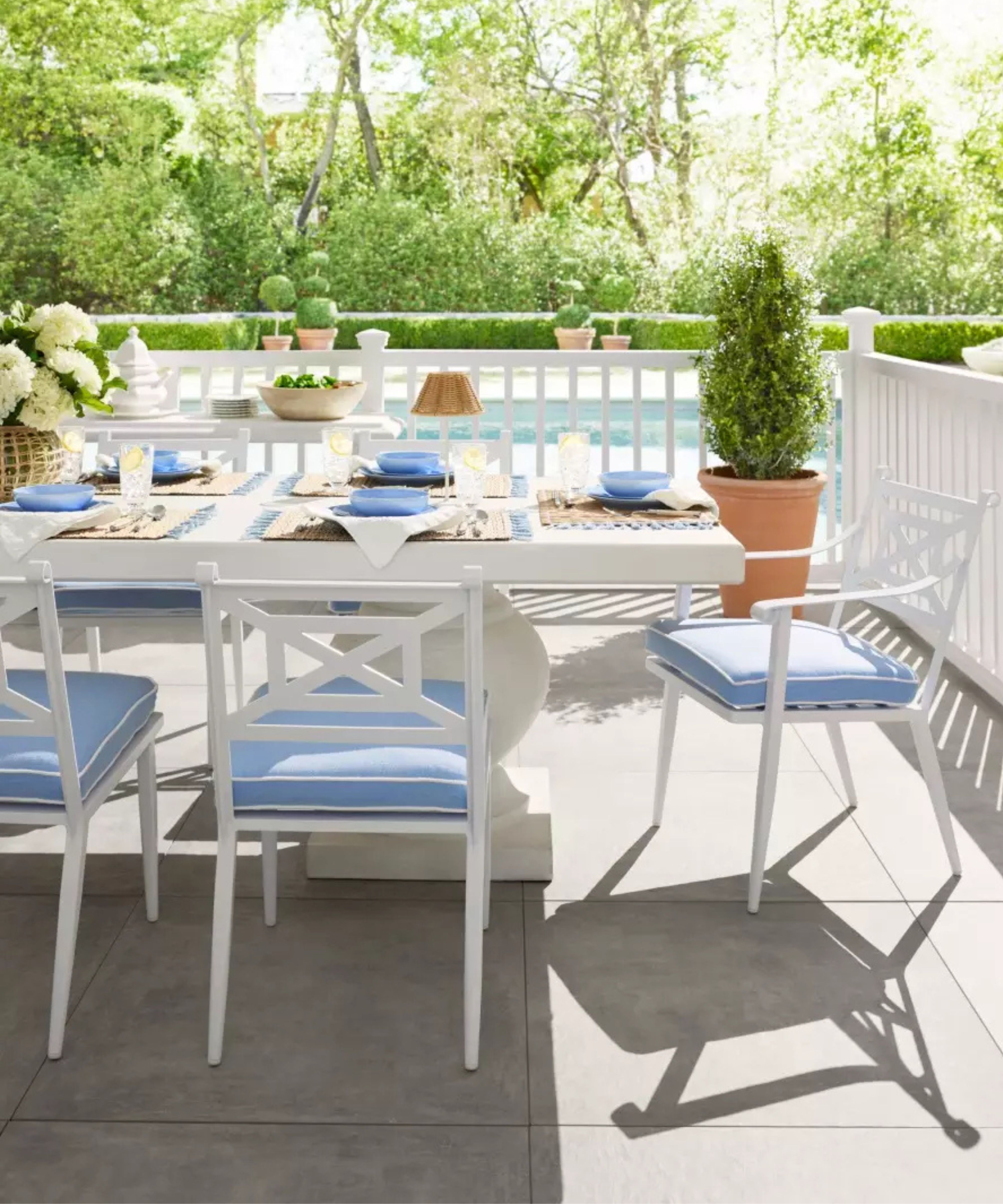 white fenced patio veranda overlooking a pool with a large white pedestal table, white chairs with blue cushions and a blue and white styled tablescape