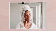 Image of a woman wearing a white dressing gown and towel around her head applying skincare, in a white frame and set against a dusky pink watercolour-style background 