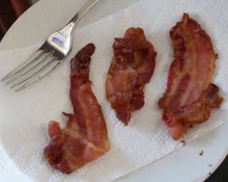 Three rashers of cooked streaky bacon made in the Gourmia 4-quart digital air fryer on paper towel and white ceramic side plate with metal fork