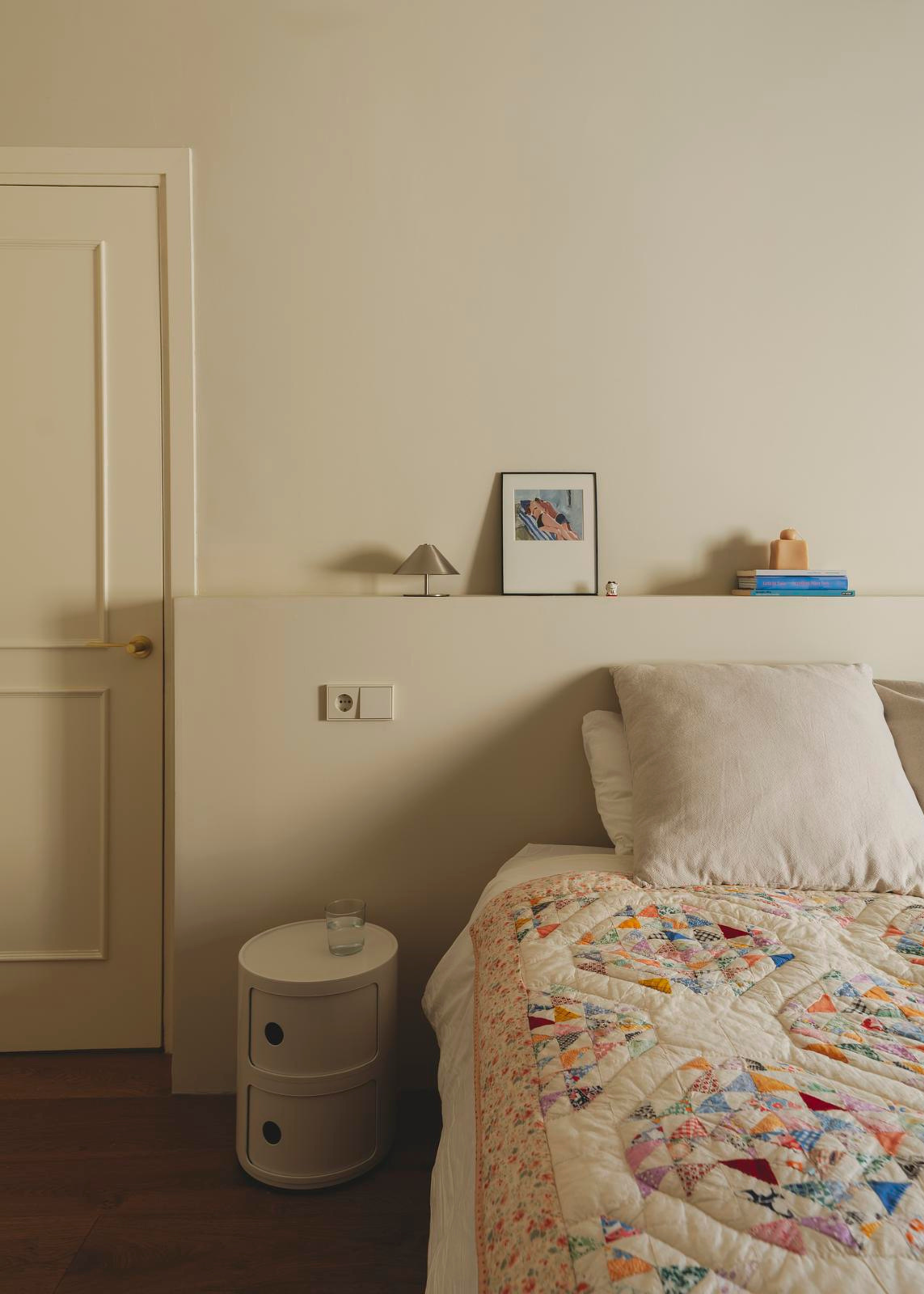 A cozy white bedroom with a bedside table, and an overhead ledge with framed art and a chrome mini lamp