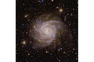 A pinkish and slightly hazy spiral galaxy in space, in front of lots of stars and more distant bright galaxies.