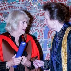 Queen Camilla and Princess Anne wearing university robes and Camilla holding a degree standing in front of a mosaic map wall