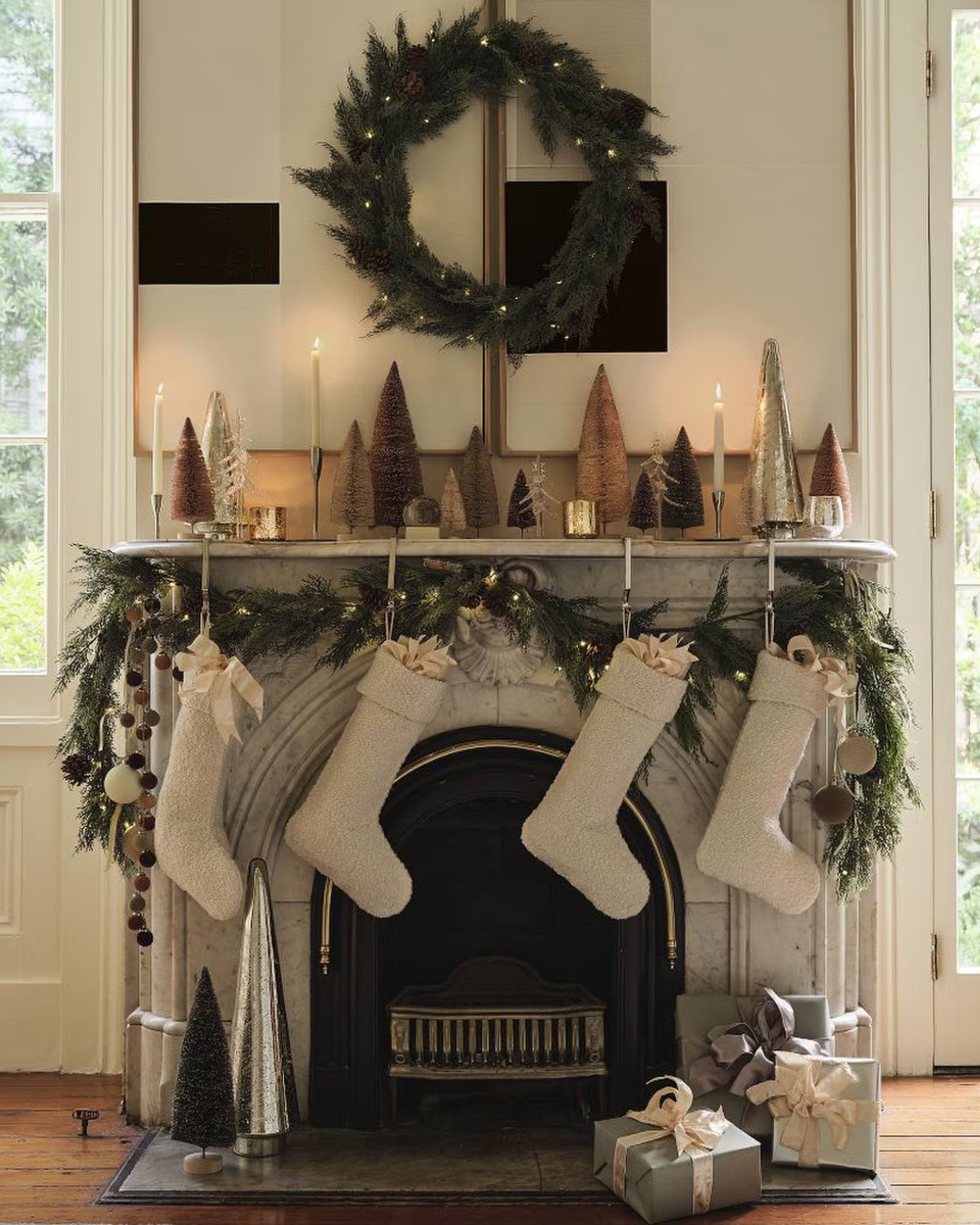 A gray stone fireplace in a white living room with a wreath hanging over it and garland on the mantel. There are four, beige boucle stockings hanging from the mantel.