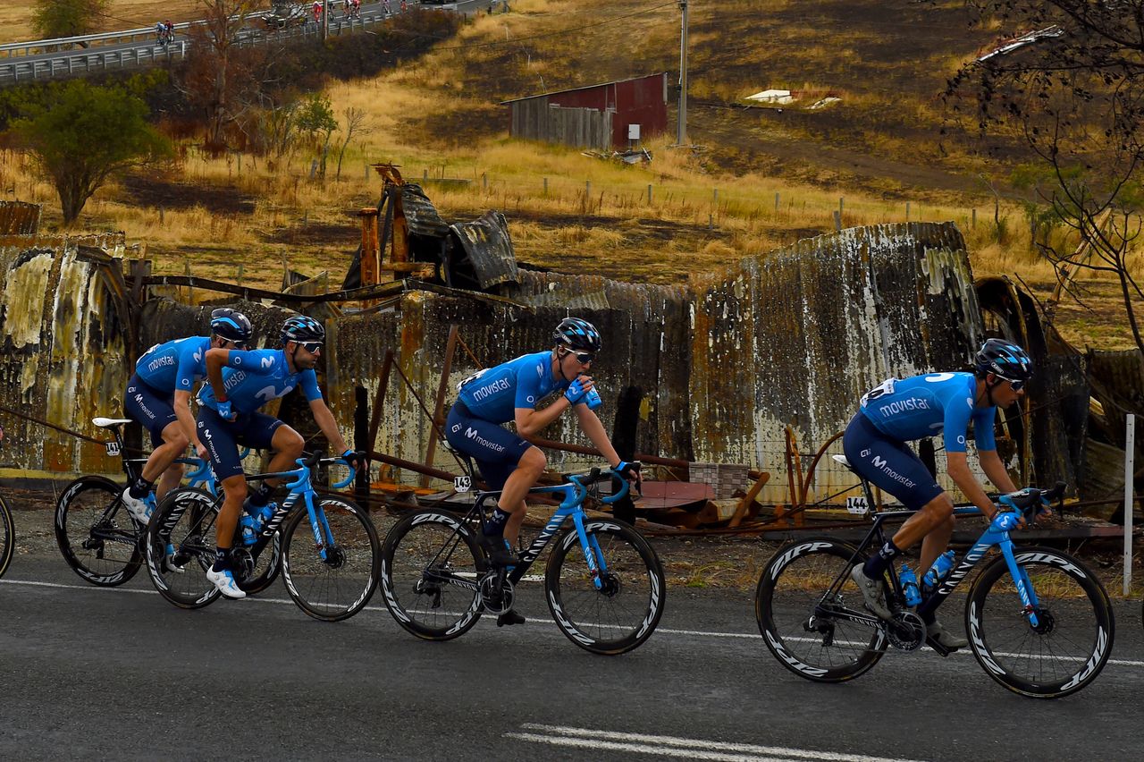 The Movistar team races past a burned-out outbuilding during stage 3 of the 2020 Tour Down Under