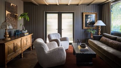 a moody brown paneled living room with low furniture, antique table and cabinet and boucle arm chairs