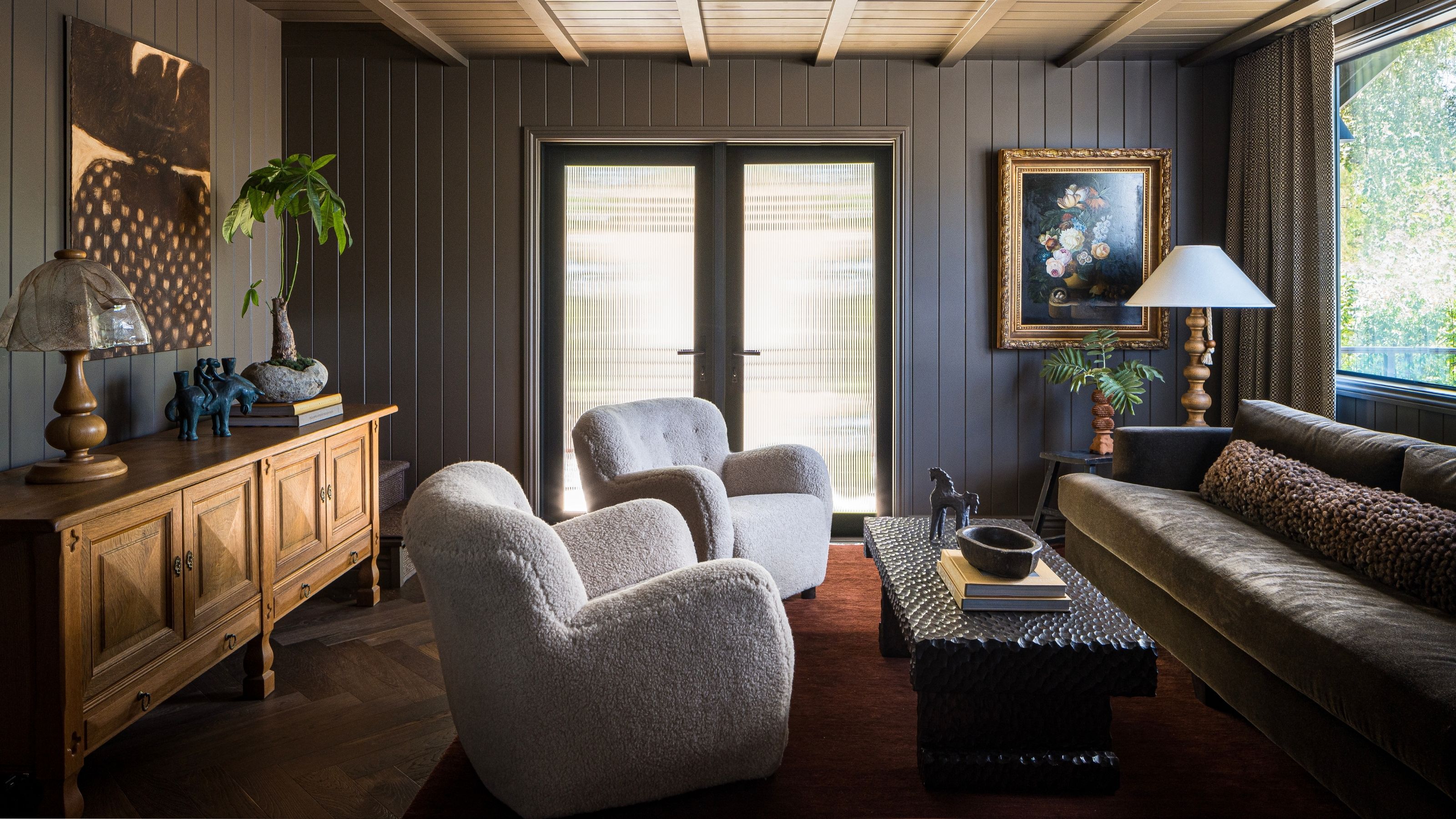 a moody brown paneled living room with low furniture, antique table and cabinet and boucle arm chairs