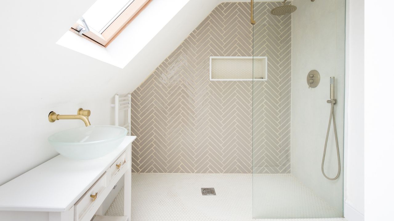 White tiled bathroom with walk in shower at far end showing taupe chevron tiling on the wall and small mosaic white tiling on the floor, and vaulted sloped ceiling with window on left