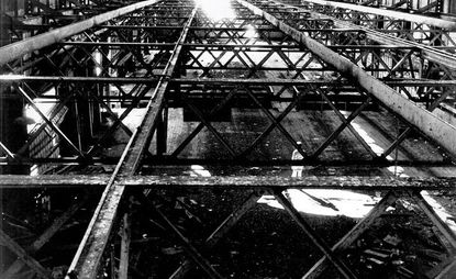 Black and white photo looking dowwards through iron grid of pier showing person dressed in white below