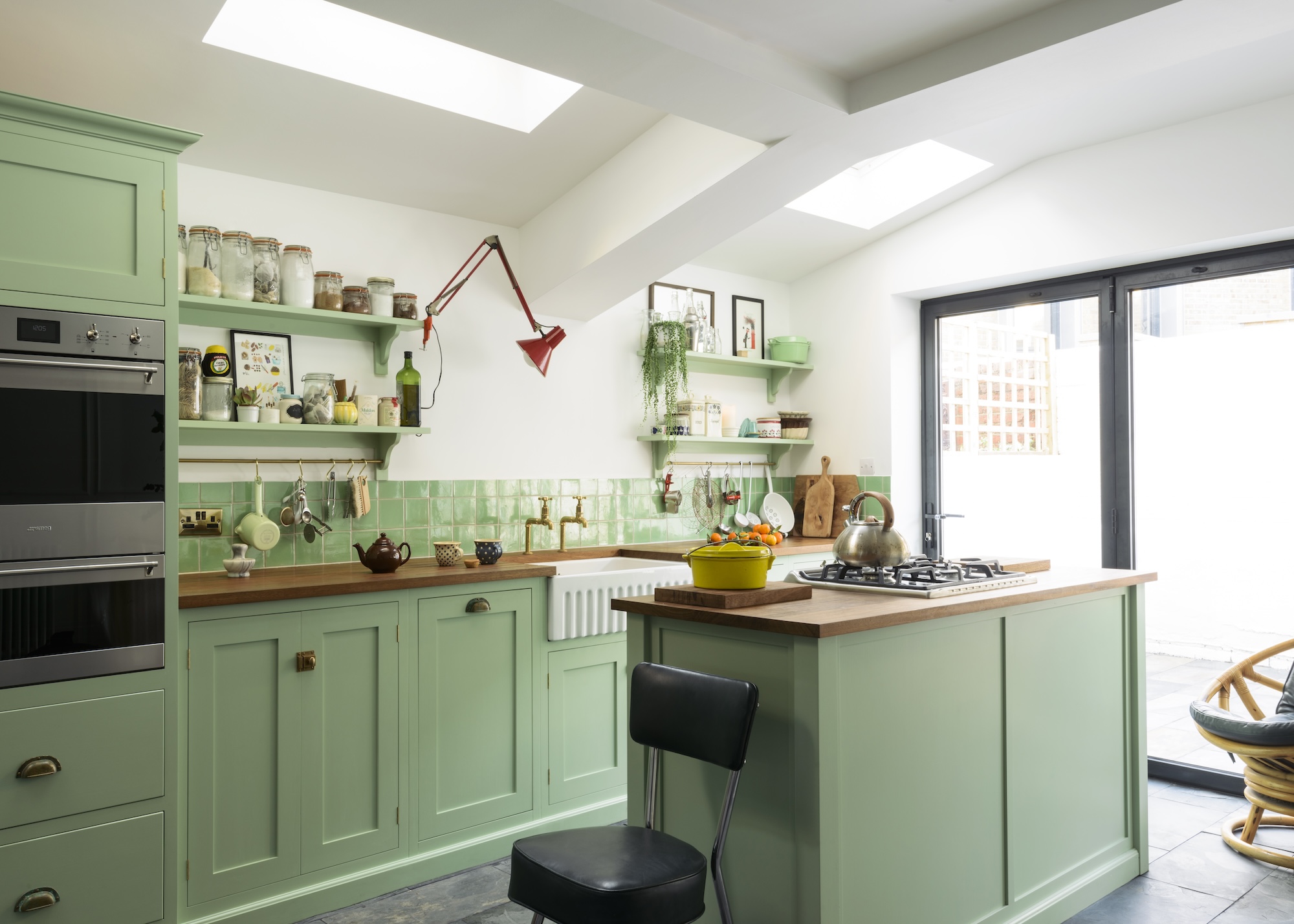 A bright green kitchen with a small island that has a stove on top of it. Beside the island is a chair. Opposite the island is a Belfast sink with open shelving above it. There is also a view of the integrated oven