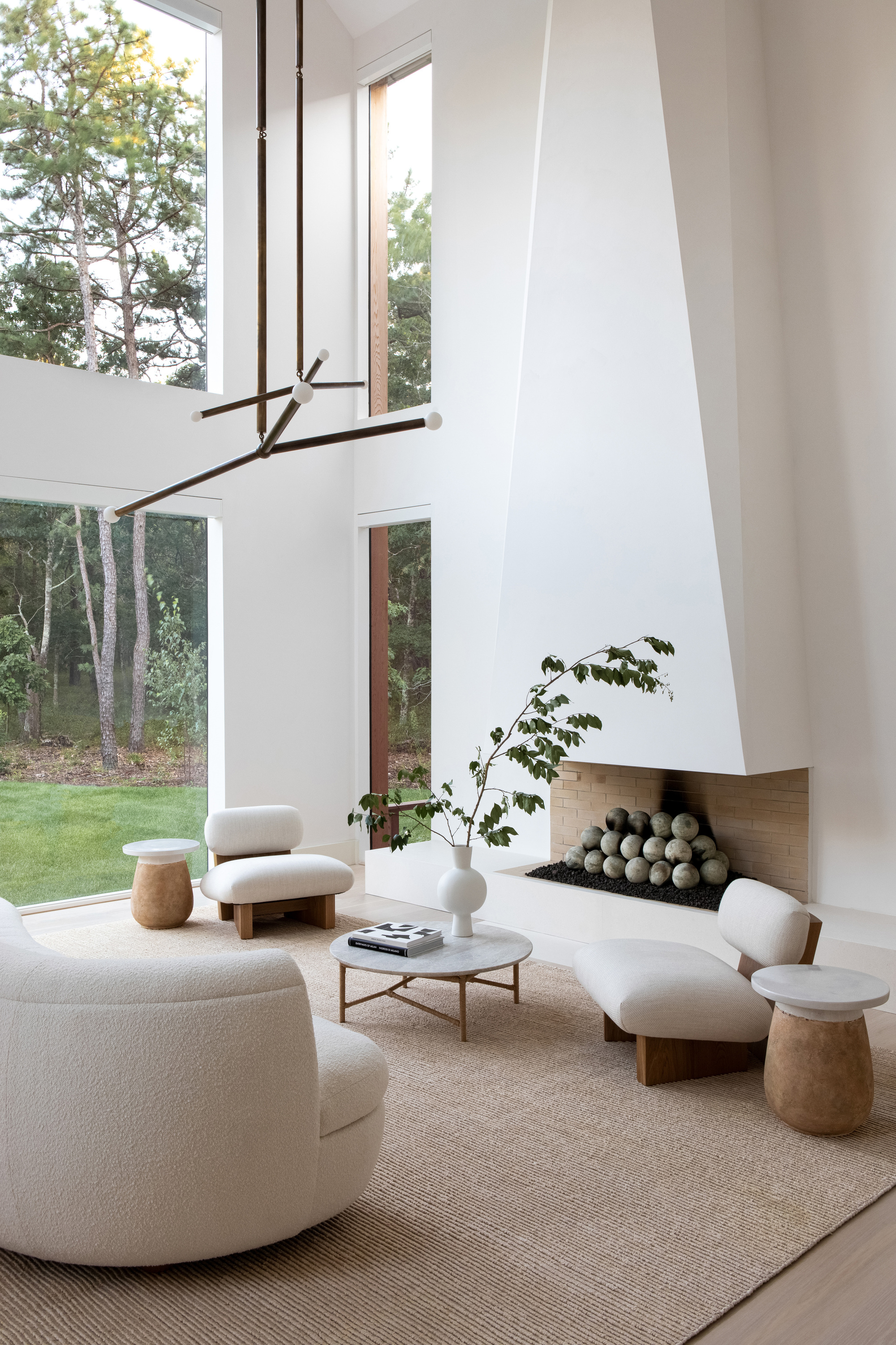 double-height beige living room idea with sculptural fireplace, beige sofa, jute rug, marble coffee table, two beige armchairs with side tables, and open doors to the garden outside