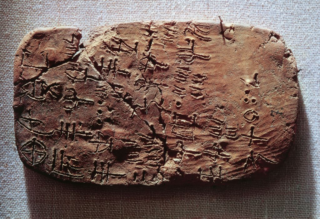 A close up of a fragmented stone tablet with small pictographs carved into its face. The reddish-brown tablet sits on a gray surface