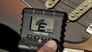 A Planet Waves guitar tuner is held in front of a Fender Stratocaster