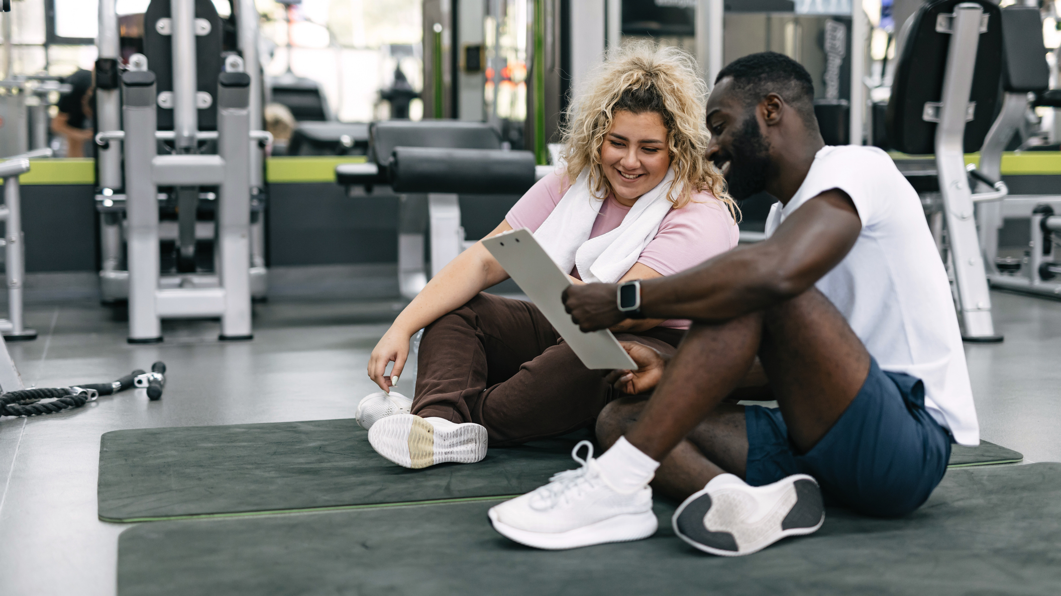 Man and woman sitting on exercise mats in gym looking at a clipboard