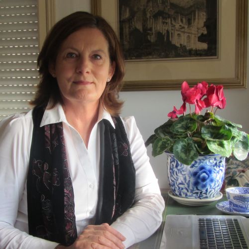 Gina Rossi, a white woman with a brown bob haircut wearing a white shirt with black velvet vest, next to a laptop and potted pink plant