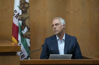 Harry Bosch (Titus Welliver) sits on the stand in a courtroom. There is a flag hanging loosely on a pole next to him, and a microphone directed into his face. He is wearing a navy blue jacket over a light blue shirt and looks very serious.
