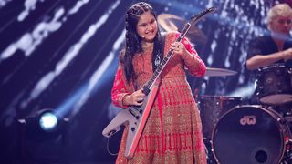 Maya Neelakantan playing a Gibson Flying V on stage, with a drummer in the background