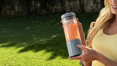 A Caucasian lady holding a small grey portable blender with a clear vessel outside. 