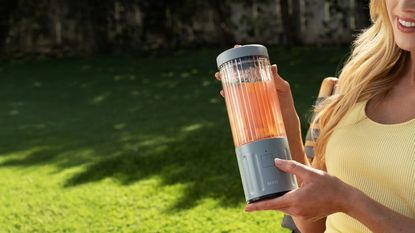 A Caucasian lady holding a small grey portable blender with a clear vessel outside.