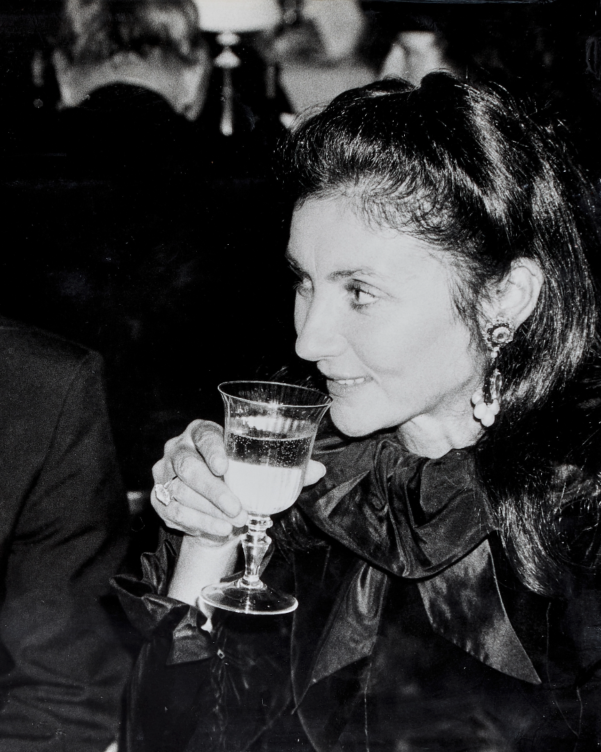 A black and white photo of Pauline Karpidas with a short bob haircut, drinking champagne wearing a black high neck top and elaborate earrings