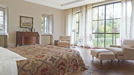A bright bedroom with large windows with sheer curtains, brown wooden floors, a large bed with a patterned quilt, matching cream upholstered chairs, and a wooden chest of drawers against the opposite wall.