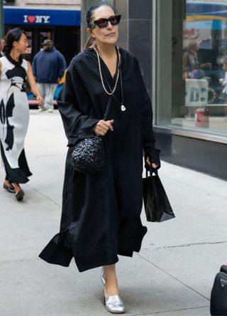 A NYFW guest wearing a black dress, silver shoes and a black bag and sunglasses outside the Melitta Baumeister show