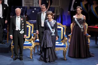 King Carl XVI Gustaf wearing a black tuxedo standing next to Queen Silvia in a blue sequin gown and diamond tiara, Crown Princess Victoria in a purple gown and diamond tiara and Prince Daniel in a tuxedo