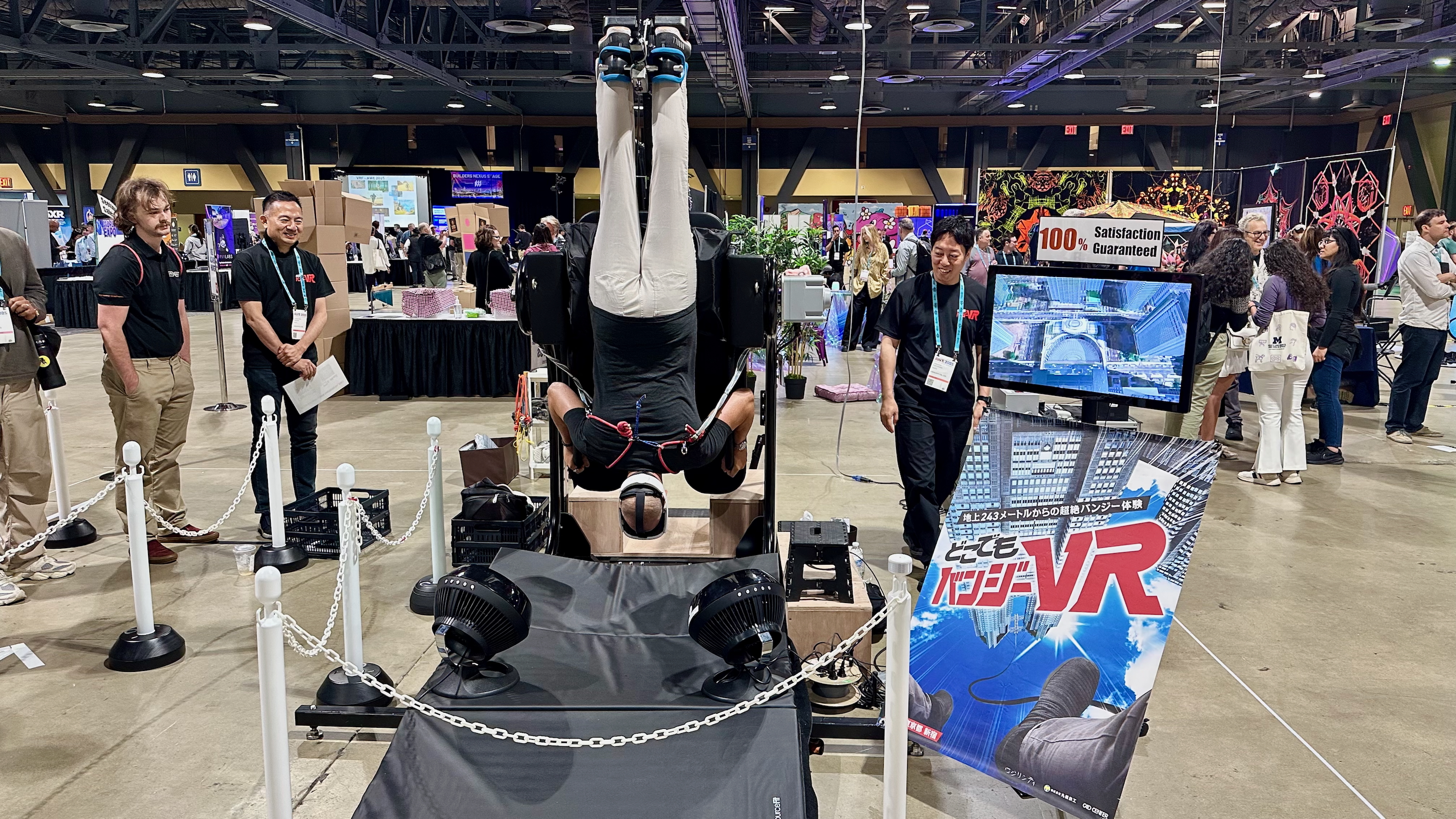 A man hanging upside-down on a contraption while wearing a Quest 3 headset on the AWE 2025 showfloor, next to a banner describing a VR bungee jumping experience in Japanese.