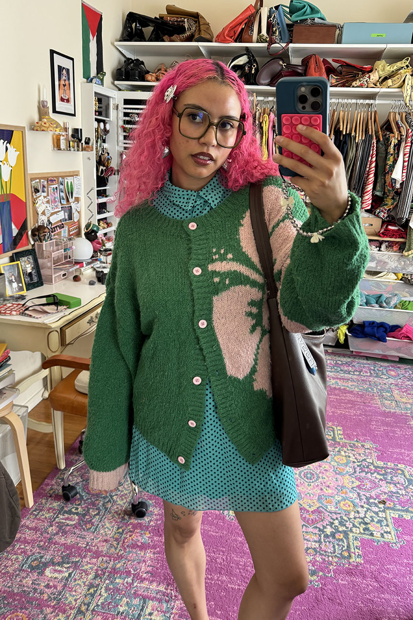 A woman with pink hair wearing a green sweater over a teal polka dot dress.