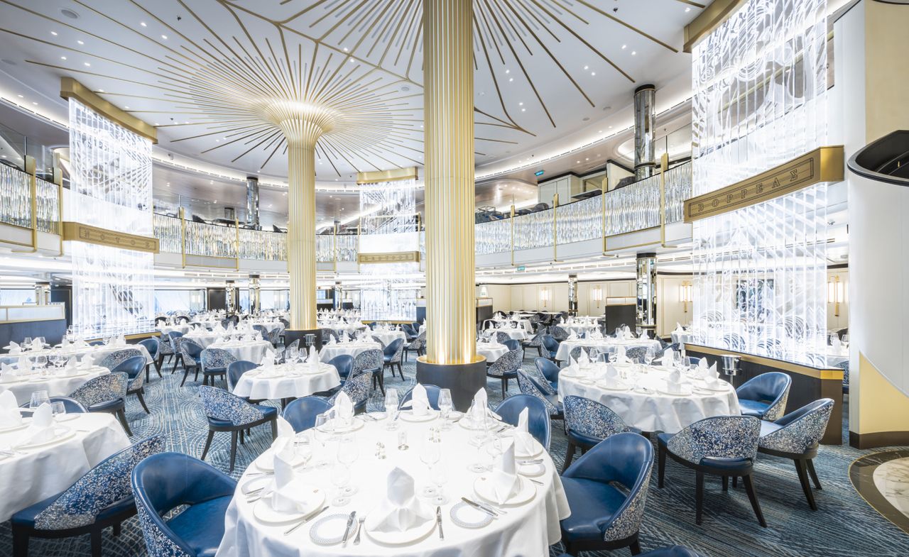 Overview of Cunard’s Queen Anne Britannia Restaurant. A series of round tables stands against a blue carpet, golden columns and glass lighting