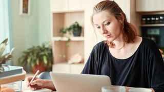 Woman using laptop and holding a pen at kitchen table