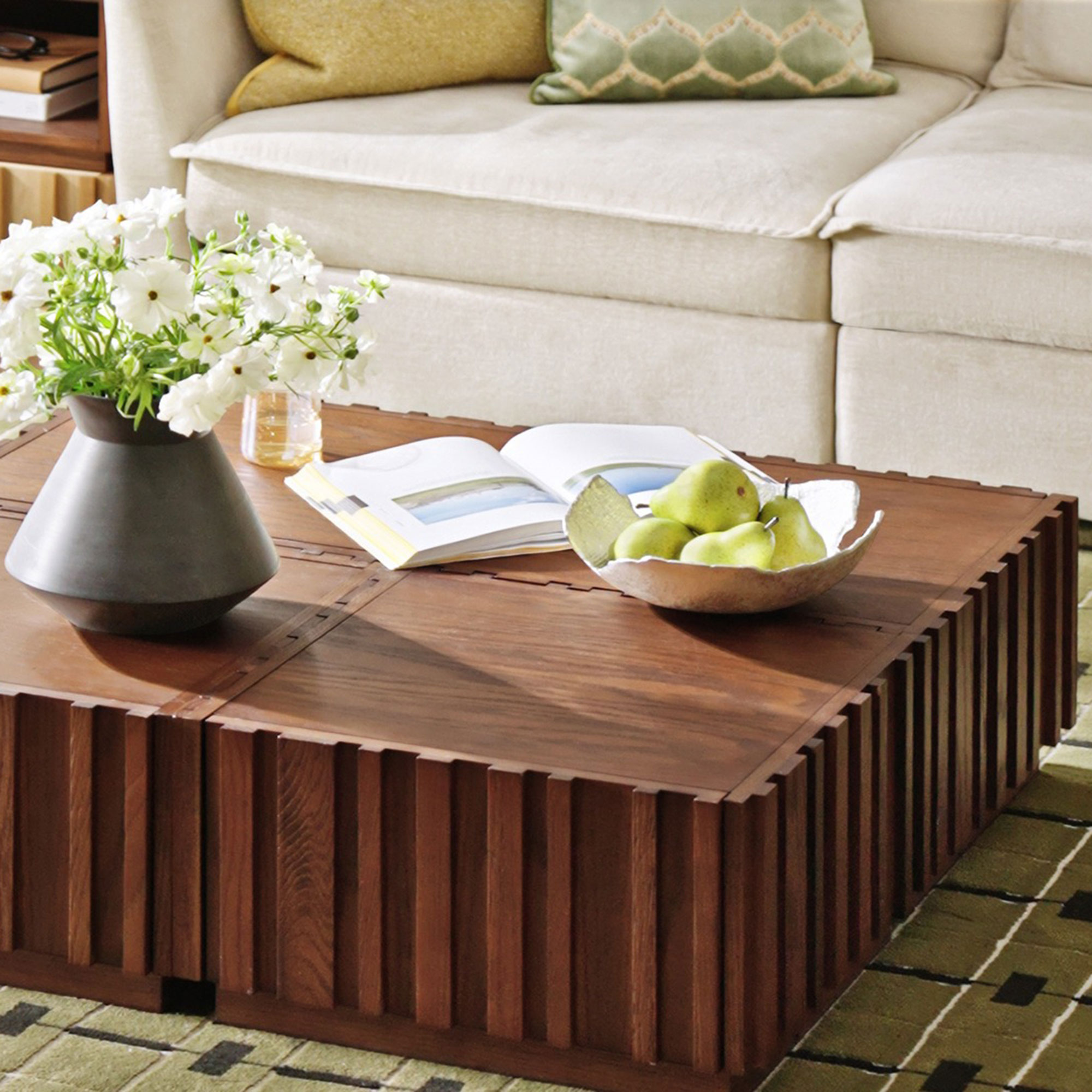 Wooden coffee table on green rug holding vase of flowers, open book and bowl of fruit