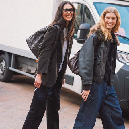 Two women are pictured walking a smiling for the camera. Each wear a jacket, dark jeans, loafers, and a leather handbag.