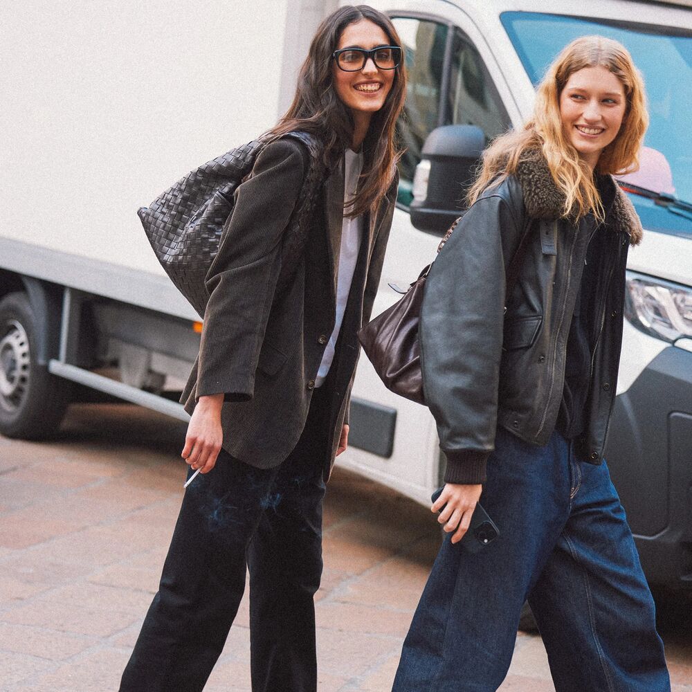 Two women are pictured walking a smiling for the camera. Each wear a jacket, dark jeans, loafers, and a leather handbag.