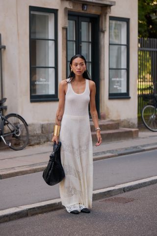 A woman wearing a maxi white knit dress with a gold cuff, gold earrings, black shoes, and a black Bottega Veneta large Jodie Bag