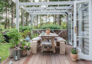 outdoor dining table on decking with wooden benches and table pergola above and trees behind
