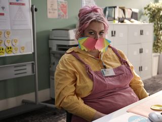 Ruby (Jayde Adams) sits at her desk in the call centre with a variety of multicolour post-it notes stuck over her mouth