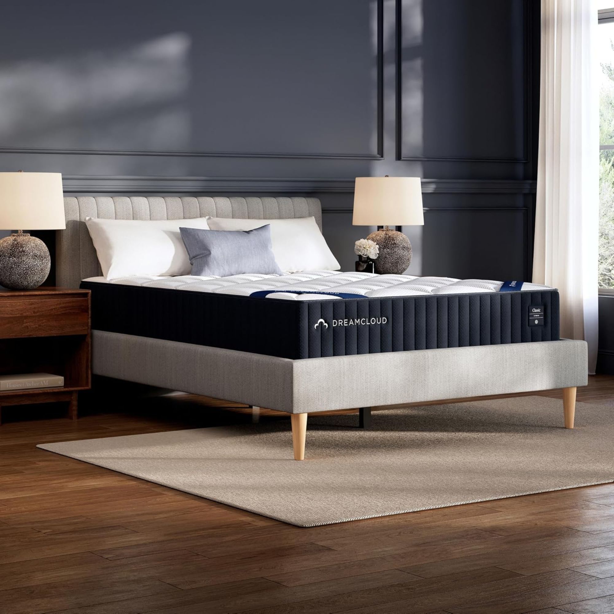 A white DreamCloud Classic Hybrid Mattress with blue edging on a grey scalloped upholstered bedframe. To either side of the bed are matching dark wood bedside tables with matching round stone lamps with white shades. The floor is dark wood, with a beige rug beneath the bed, and the walls are dark blue and panelled. To the right is a large window with an open sheer white curtain.