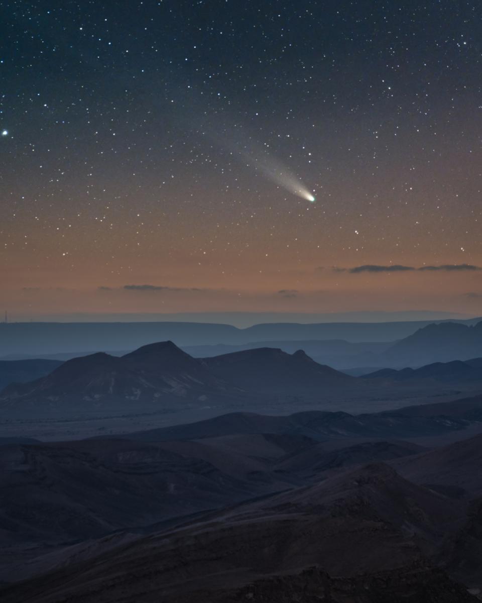 The shadowing layers of overlapping mountains in the rocky Israeli desert sit below a sunset painted sky blotted with stars. Above the mountains, a bright comet with a long, fading tail floats amongst the star splattering.