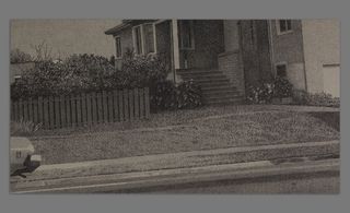 Black and white image shows the front of a house with a garden fence