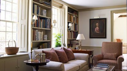 A cozy living room with build in book shelves between windows. A cream sofa in front beside a brown upholstered arm chair.