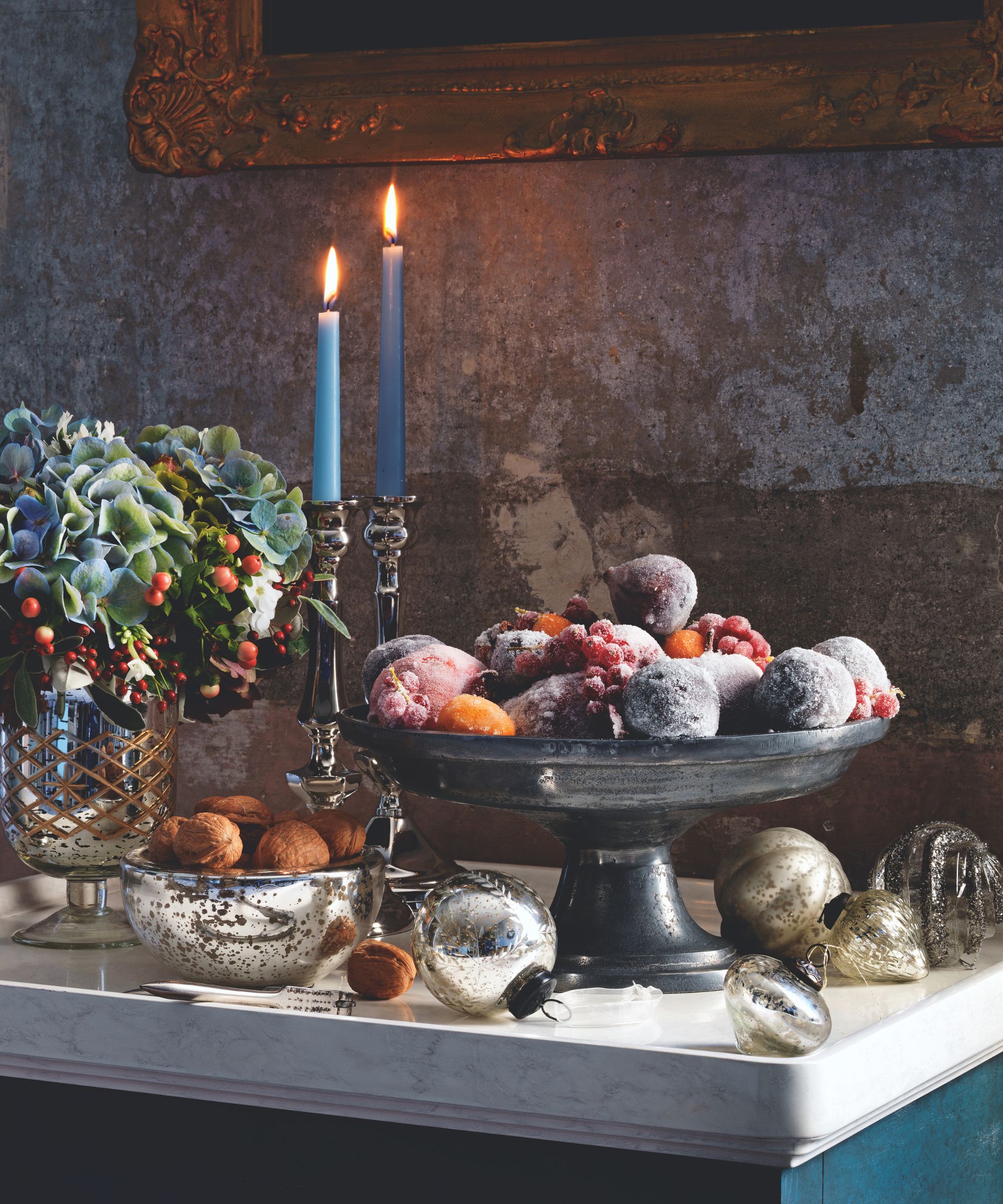Frosted fruit and nuts in bowls on a counter with candles and Christmas decorations