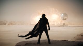 Still from the movie Dune: Part 2. Paul Atreides is standing in the foreground, his cloak billowing out behind him in the wind. Far off in the distance. In between there is a large, floating shiny sphere.