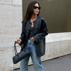 Woman wearing collarless leather jacket with jeans and black handbag, walking on street.