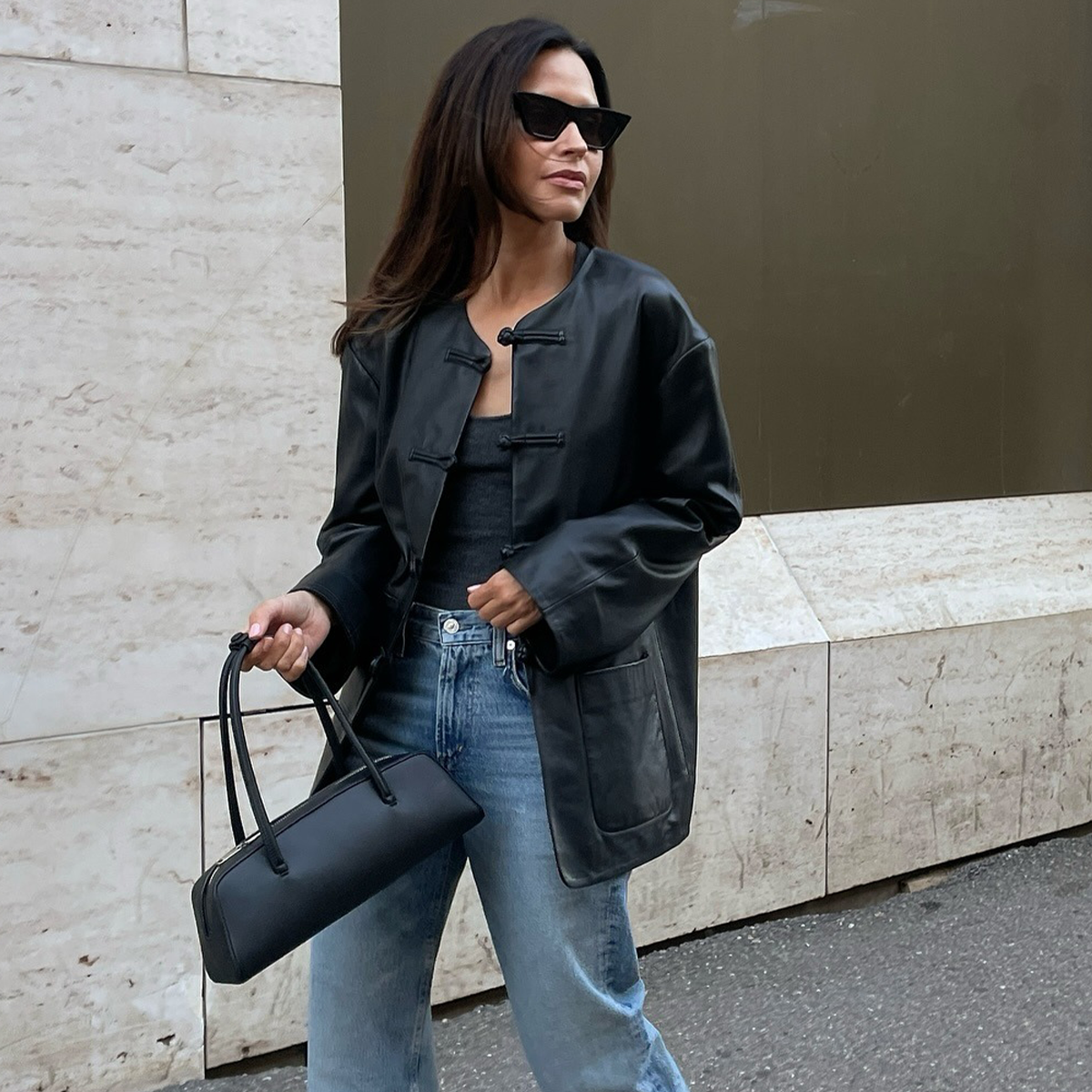 Woman wearing collarless leather jacket with jeans and black handbag, walking on street.
