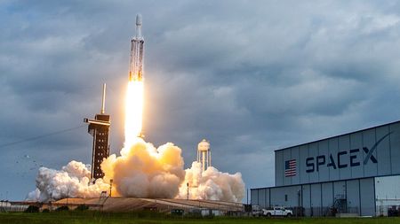 a triple-core rocket blasts off under a sunny, partly cloudy blue sky. smoke expands below next to an obscured launch tower, near a long white hanger with SPACEX written on the side. To the left, a bird flies away.