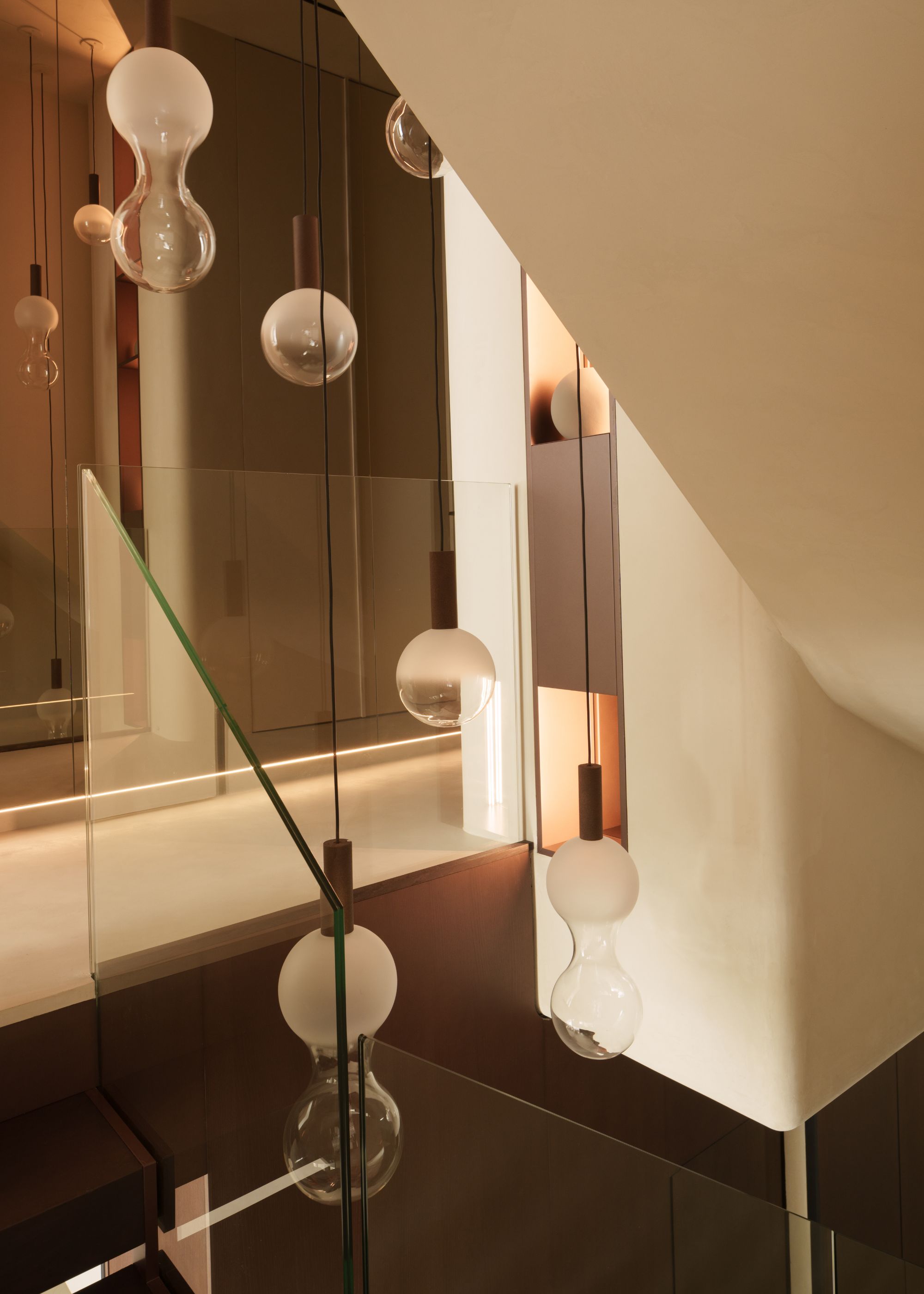 Stairway pendant lighting that has a series of glass hourglass-shaped shades that are frosted at the top, hung at varying lengths on black string in a neutral walled space with wood details