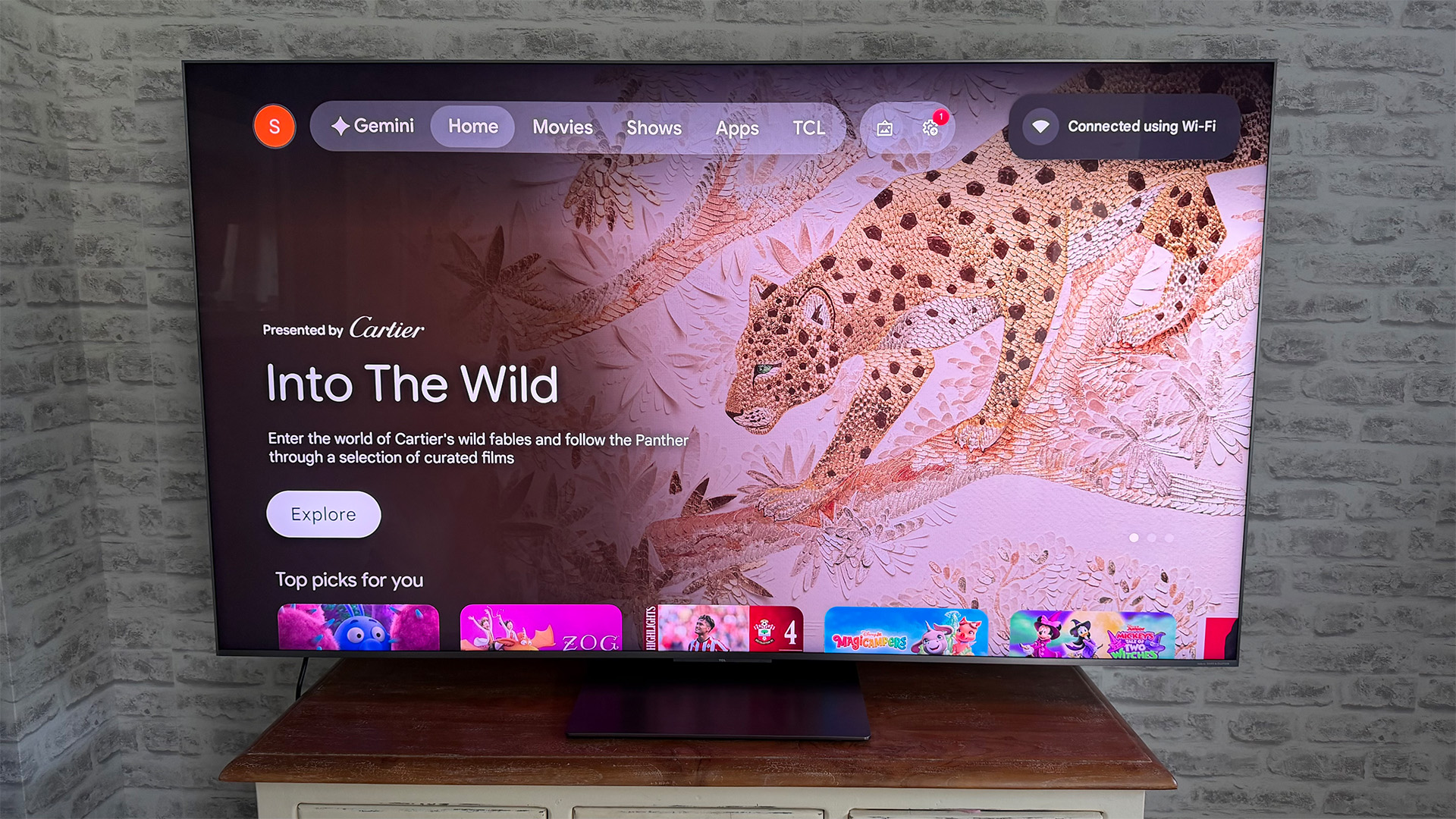 TCL C8L Mini LED TV in front of brick wall on screen is Google TV home page