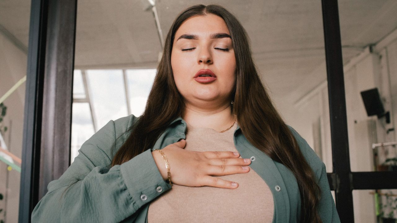 A woman in an office building practices breathwork with her eyes closed and a hand on her chest.