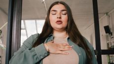 A woman in an office building practices breathwork with her eyes closed and a hand on her chest.