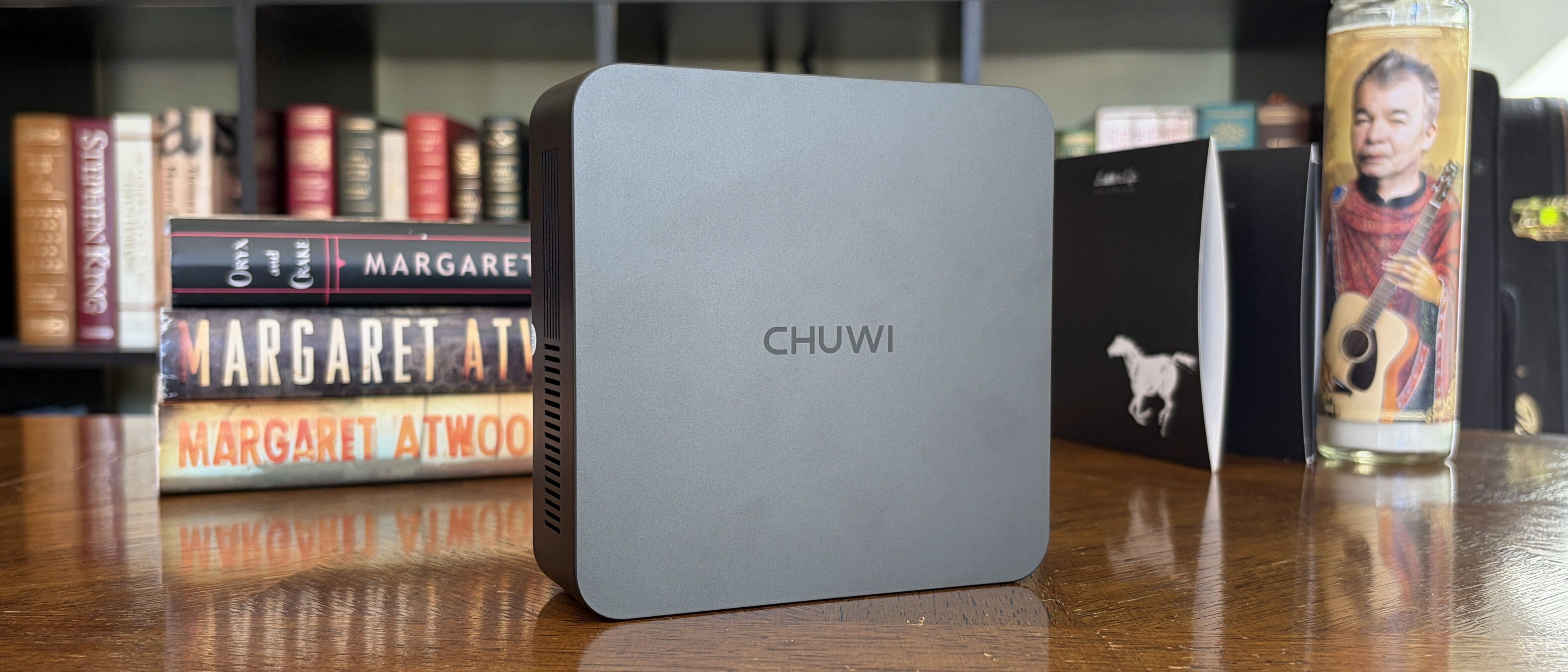Black CHUWI mini PC on a wooden surface, surrounded by books and a decorative candle featuring a man with a guitar.